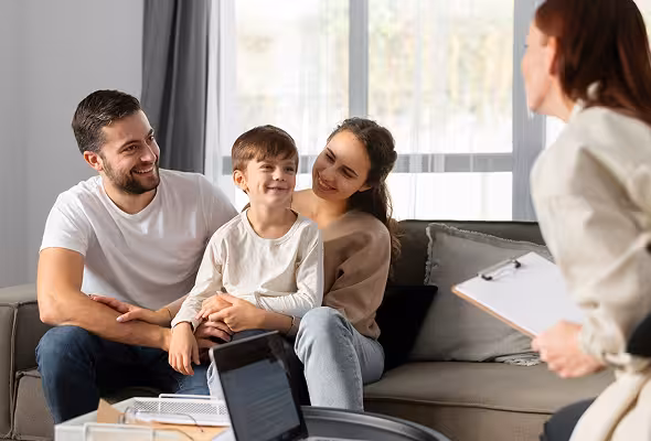 A smiling family of three sitting on a couch, talking with a professional holding a clipboard.