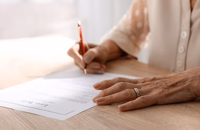 Close-up of an elderly person signing a legal document on a wooden table.