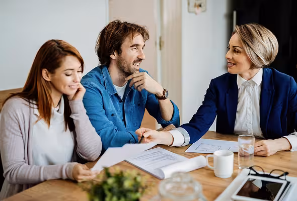 Three professionals engaged in a discussion around a wooden table with documents and electronic devices.