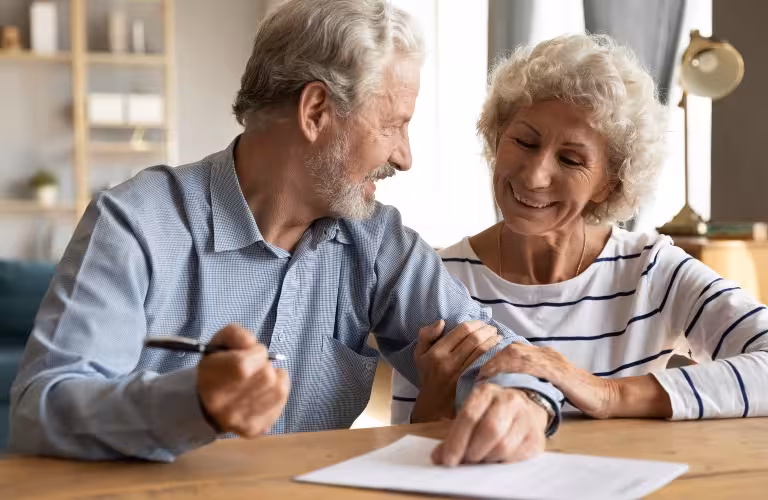 Elderly couple smiling and reviewing documents together at a wooden table.