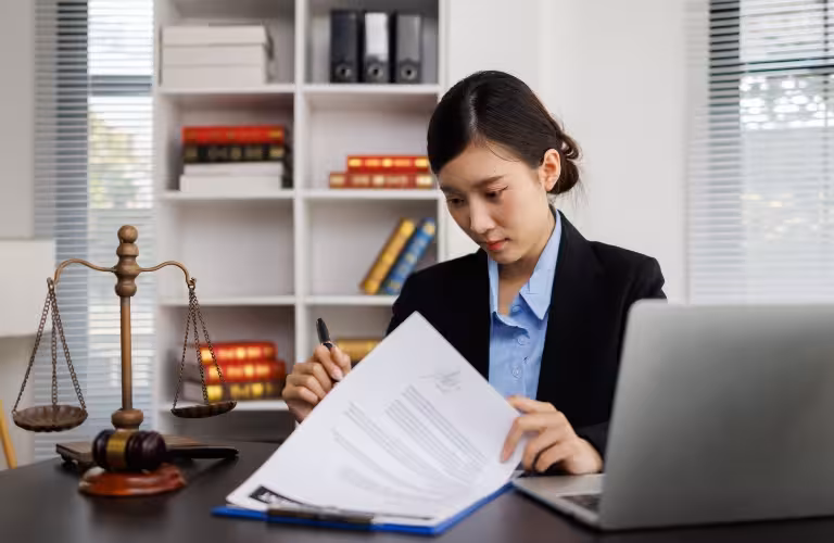 Professional woman in formal attire reviewing documents at desk with legal scales and laptop.