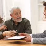 An older couple reviews and signs a document handed to them by an advisor at a wooden table_s