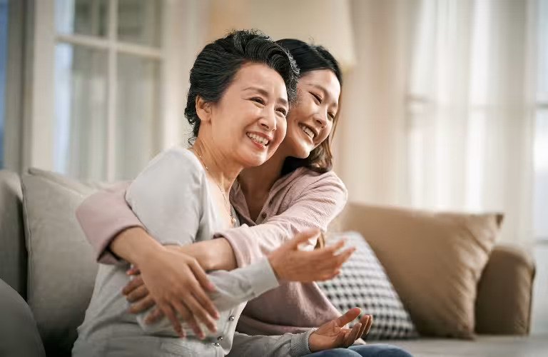 Two women, one older and one younger, smiling and embracing on a beige sofa in a bright room.
