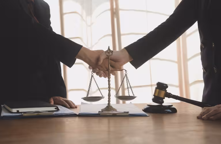 Two individuals in dark suits shaking hands over a desk with scales of justice and a gavel.